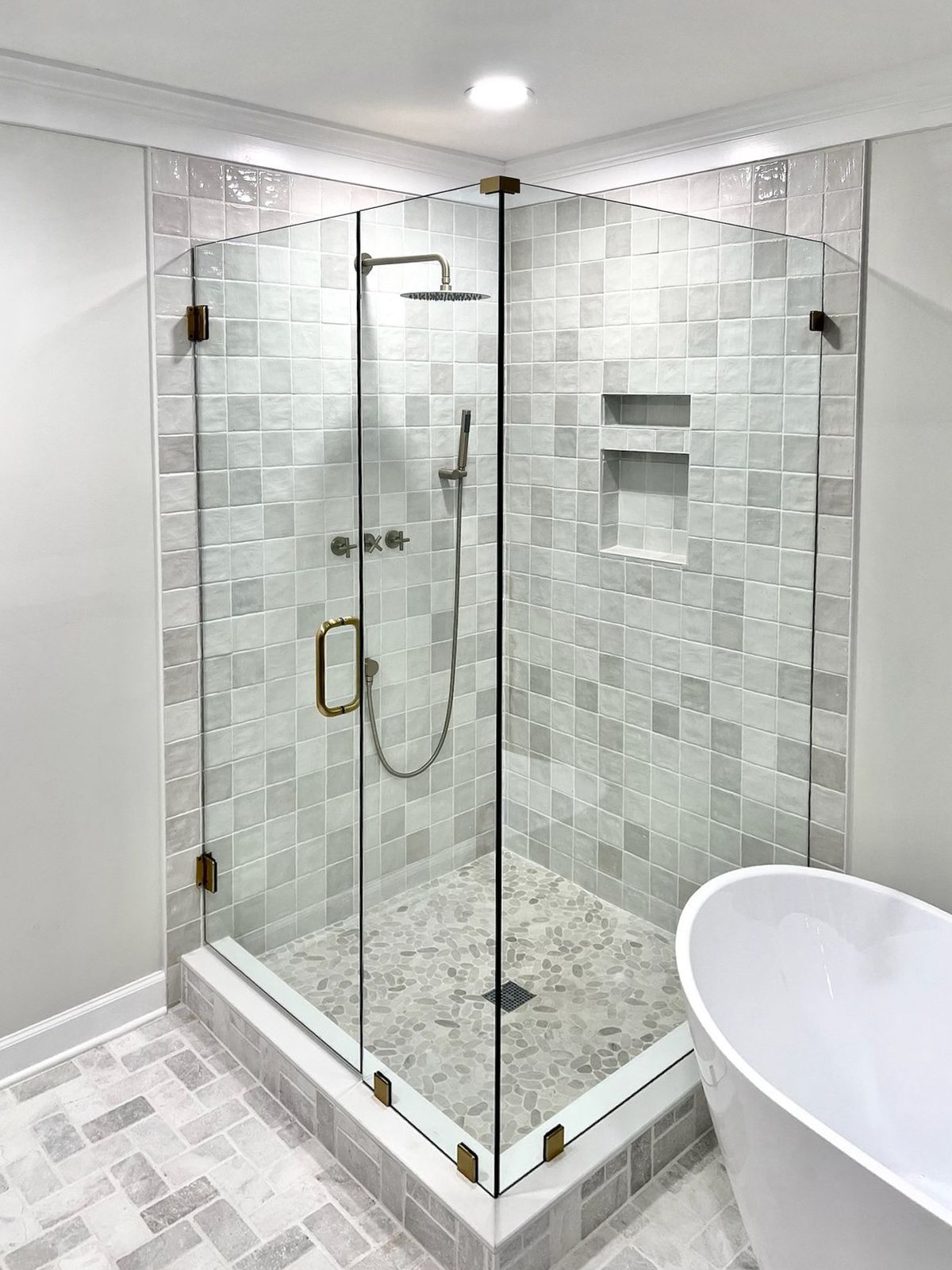 Frameless corner shower with brushed brass handle and hinges, next to a freestanding tub