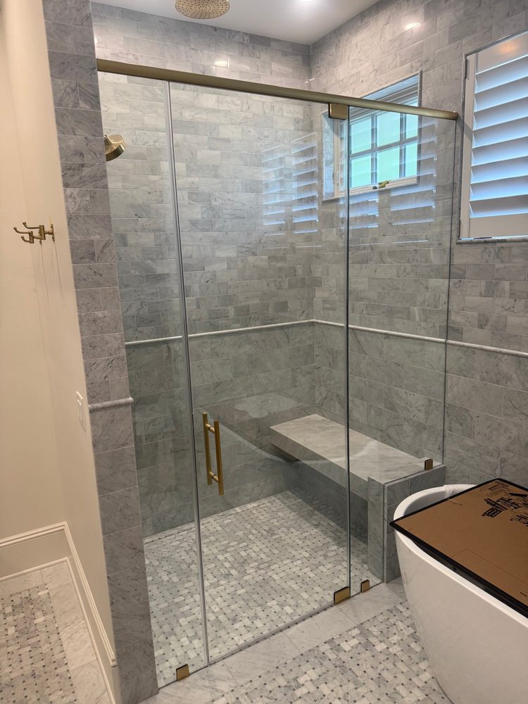 Frameless glass shower with a brushed brass header bar and a marble bench, next to a freestanding tub