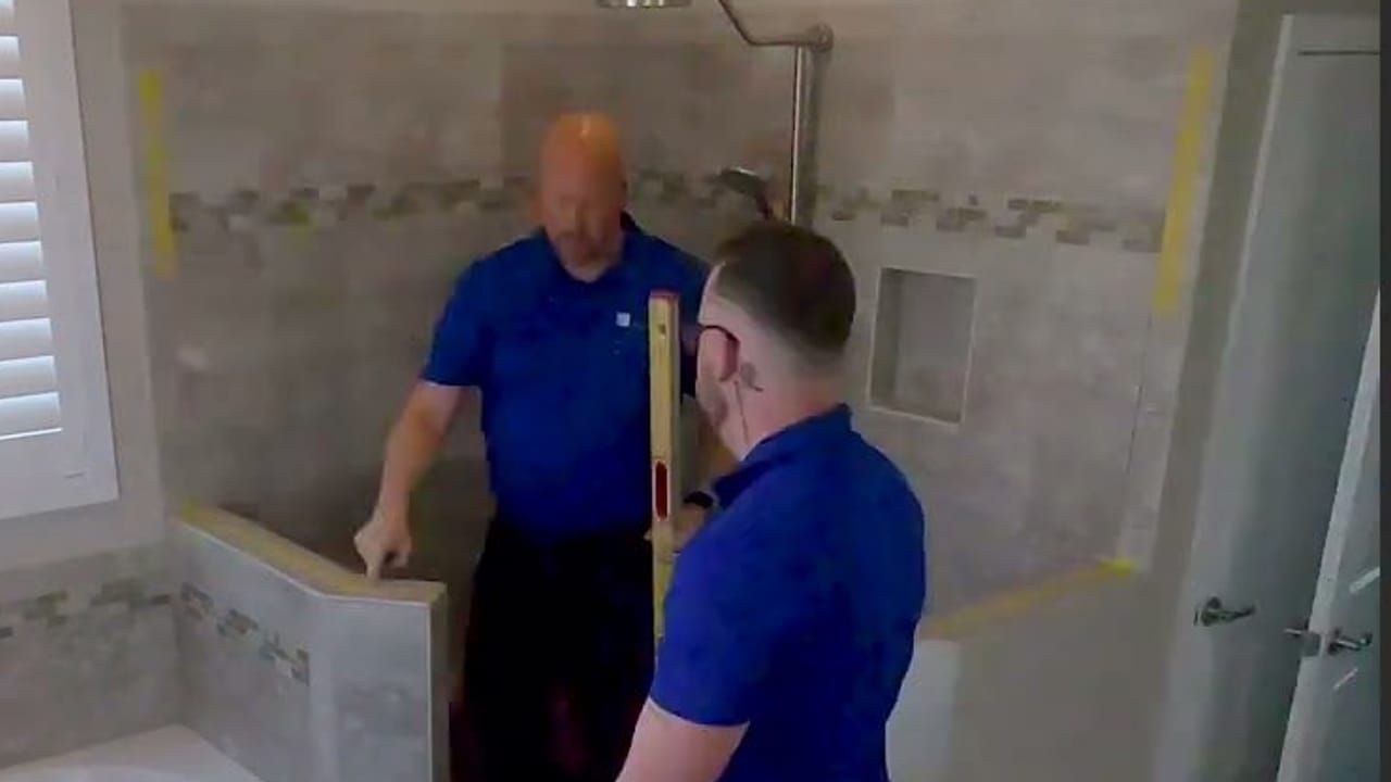 Two Shower Glass installers setting a frameless glass panel on a tiled knee wall